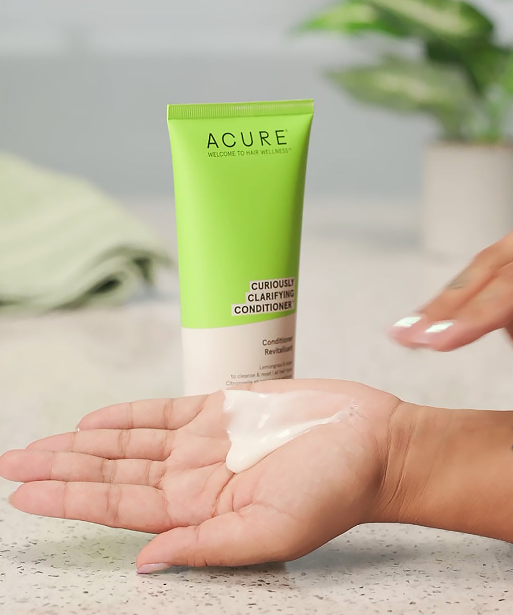 Acure Curiously Clarifying Conditioner tube with cream on a hand against a neutral background