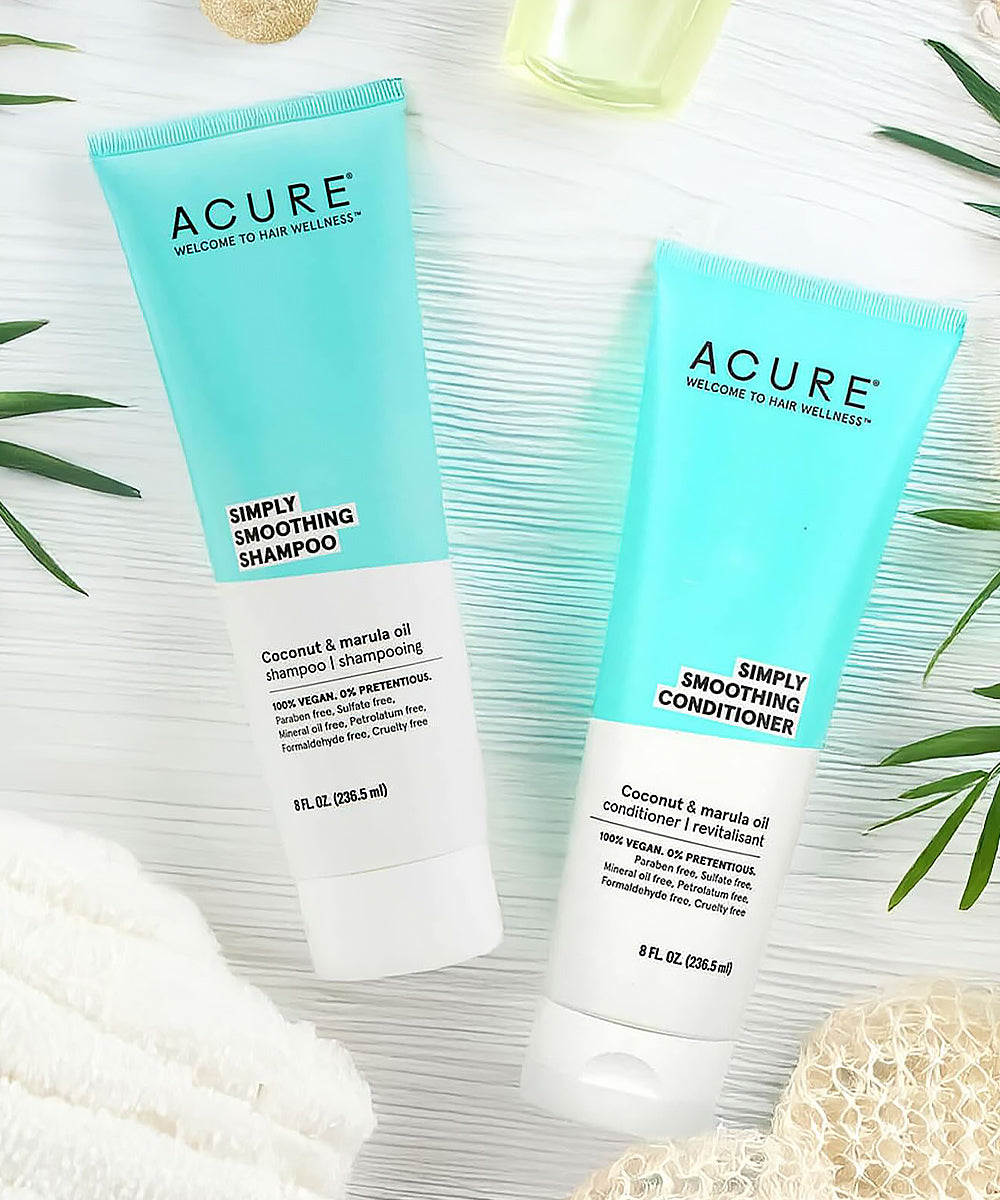 Acure Simply Smoothing Shampoo and Conditioner on a textured surface with green leaves.