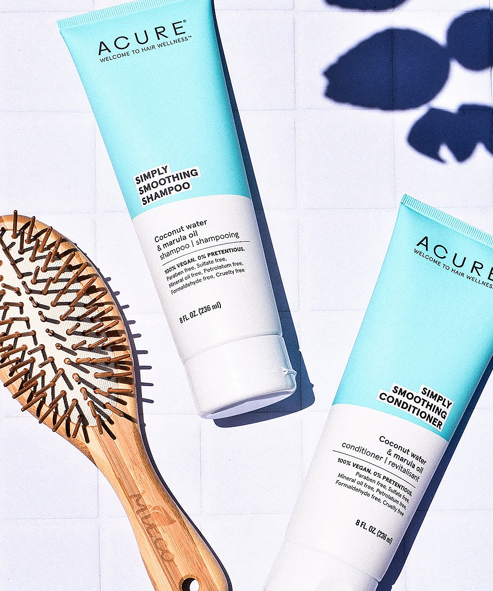 Acure Simply Smoothing Shampoo and Conditioner bottles with a wooden hairbrush on a light background