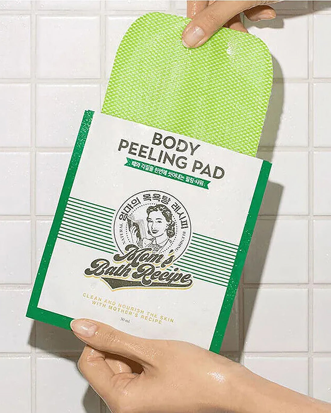 Body peeling pad packaging held by a hand against a tiled bathroom background