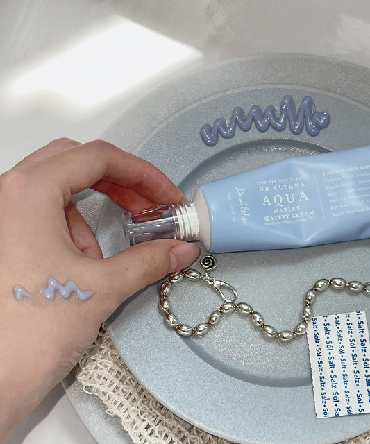 Hand holding a small bottle over a tube of cream on a textured surface with a chain and tag.