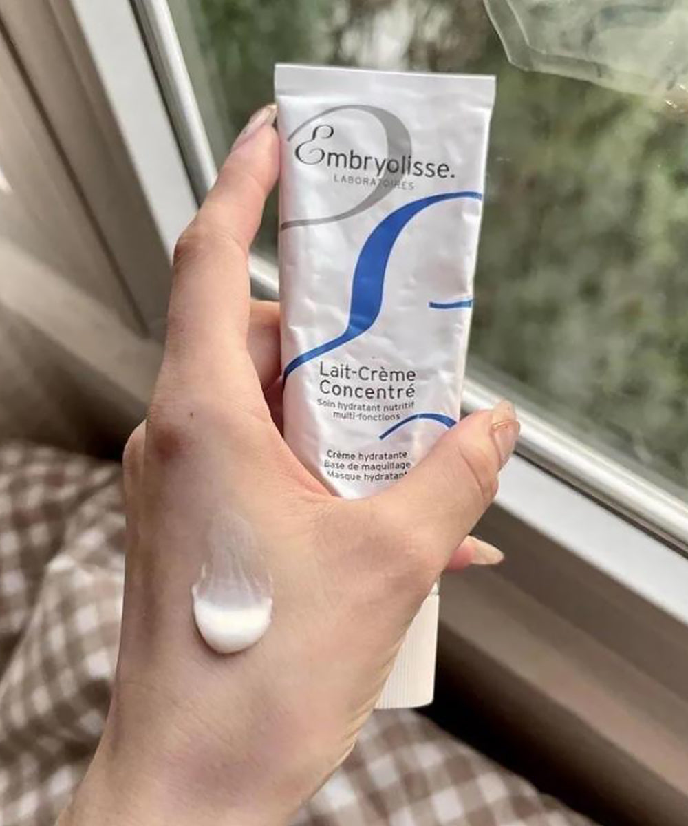 Hand holding a tube of Embryolisse Lait-Crème Concentré with a blurred window background