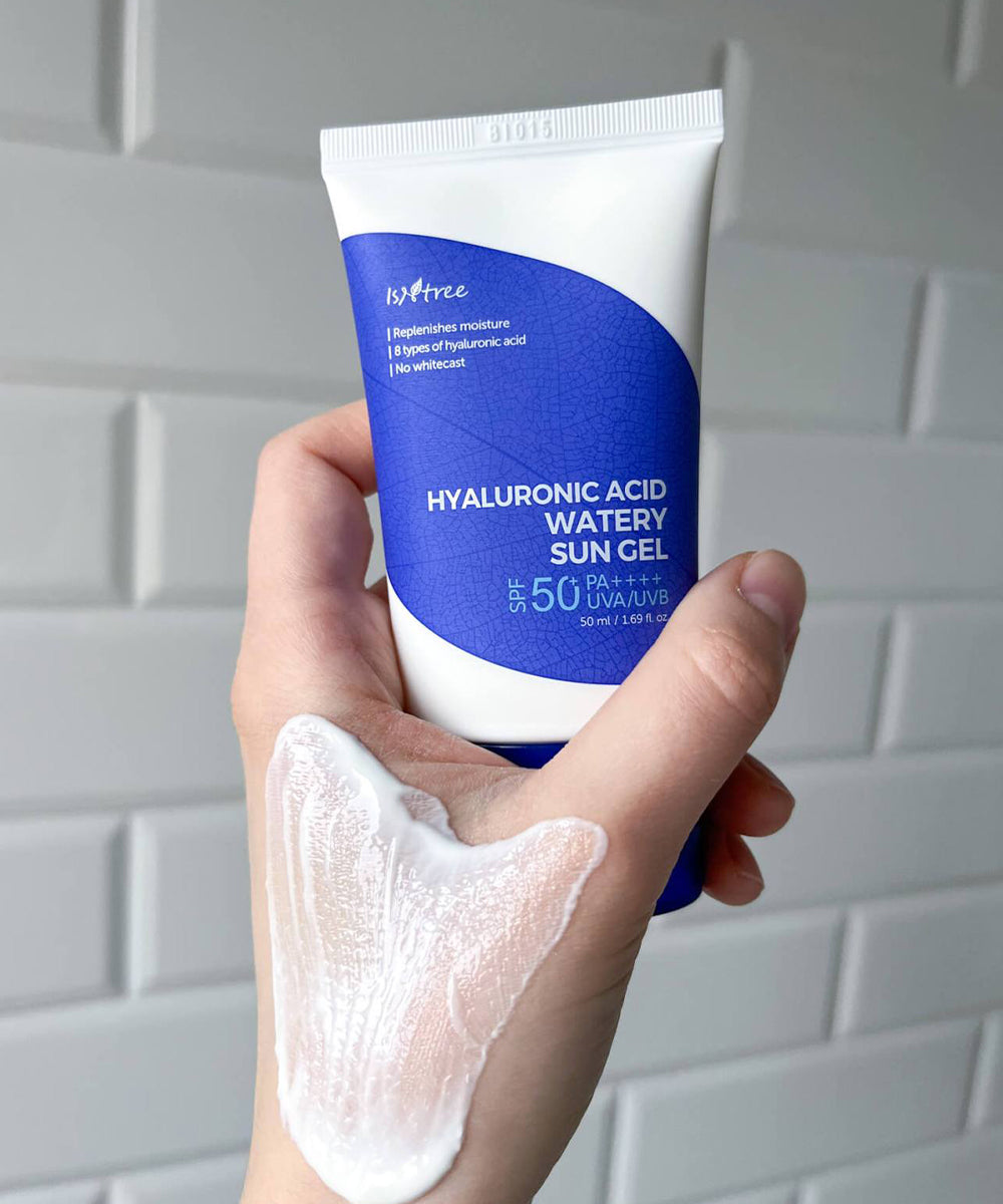 Hand holding a tube of hyaluronic acid sun gel with a visible application on the palm against a tiled wall background.