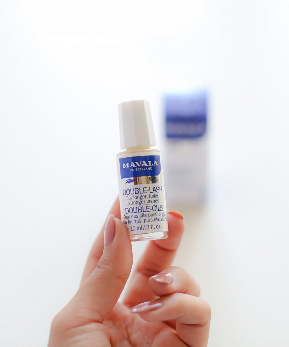Hand holding a Mavala Double Lash oil bottle against a blurred background