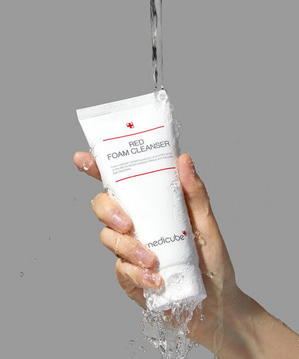Hand holding a tube of Red Foam Cleanser with water being poured over it against a gray background