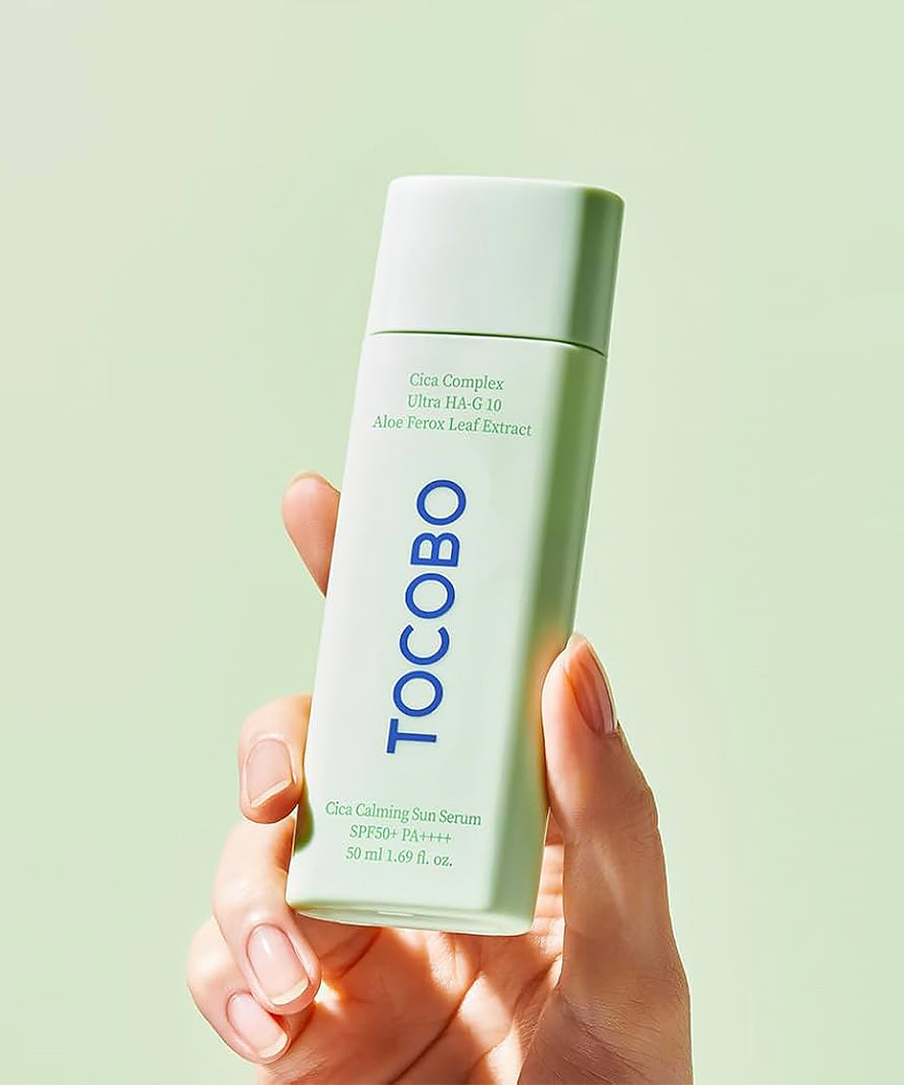 Hand holding a bottle of Tocobo skincare serum against a light green background