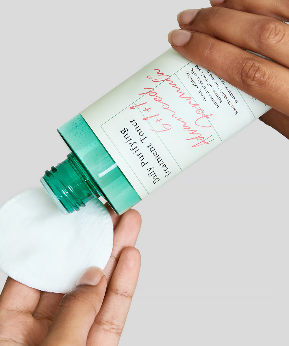 Person holding a skincare product with a green cap and white container on a light gray background