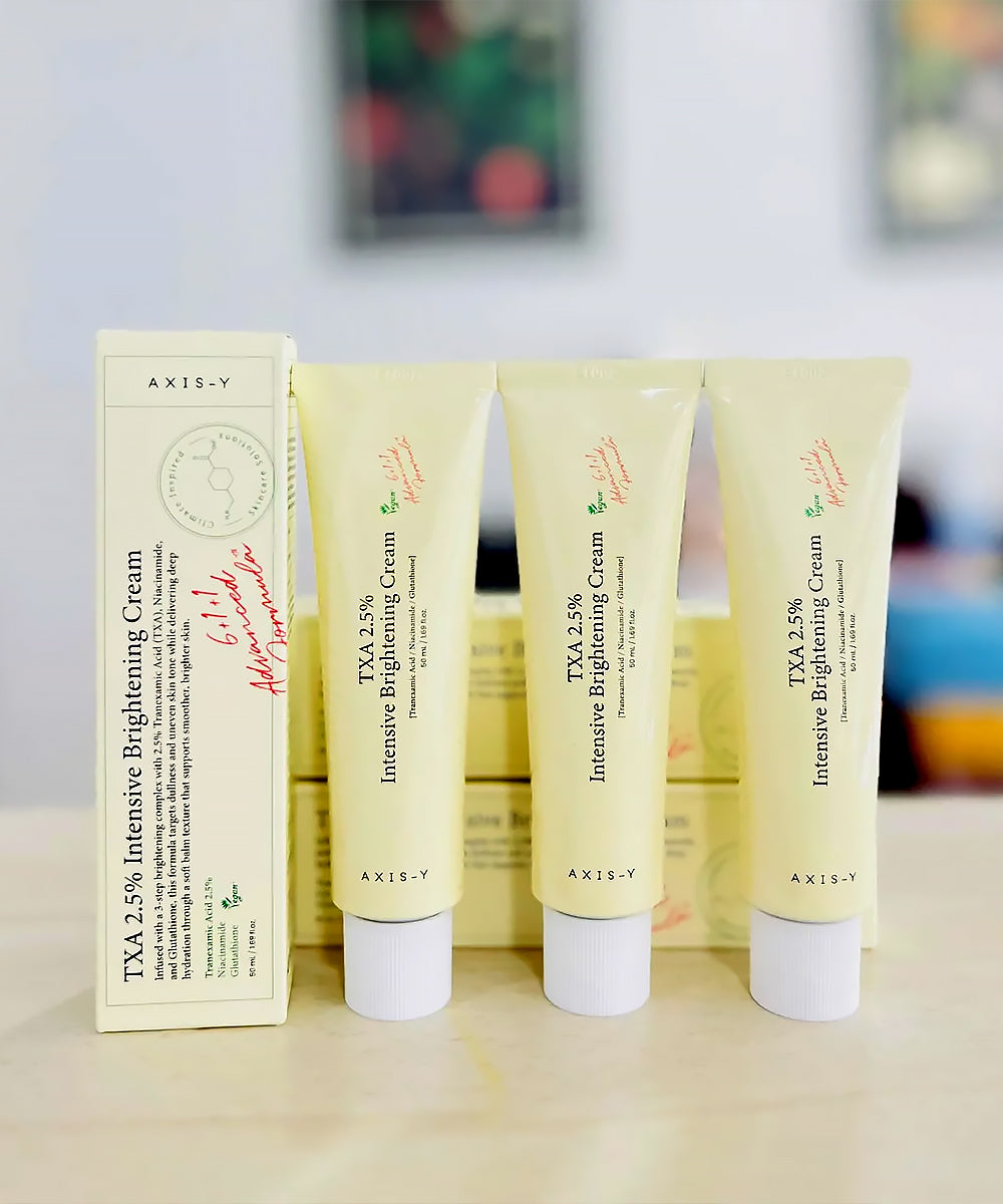 Three tubes of AXIS-V TXA 2.5% Intensive Brightening Cream on a wooden surface with a blurred background.