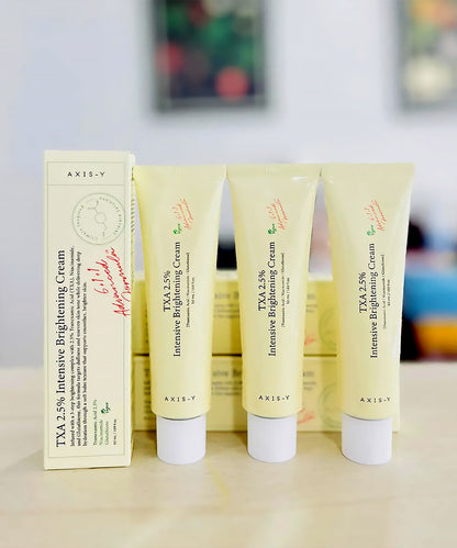 Three tubes of AXIS-V TXA 2.5% Intensive Brightening Cream on a wooden surface with a blurred background.