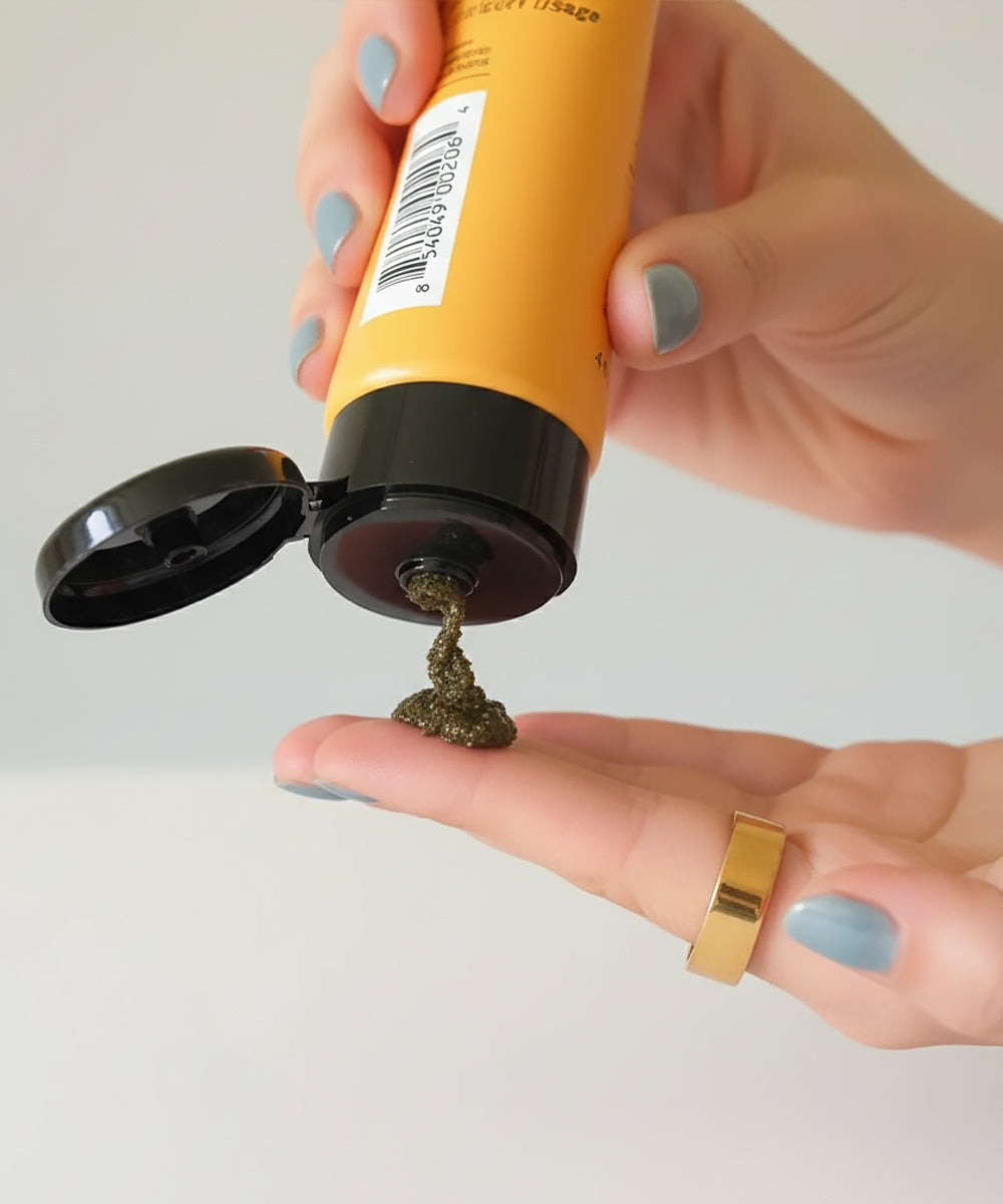 Hand holding a yellow tube with black cap, dispensing a small amount of greenish substance onto a palm.