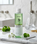 A lifestyle display of the Anua Clarifying Toner on a white marble counter, accompanied by fresh green botanicals and a glass beaker, highlighting its clean beauty aesthetic