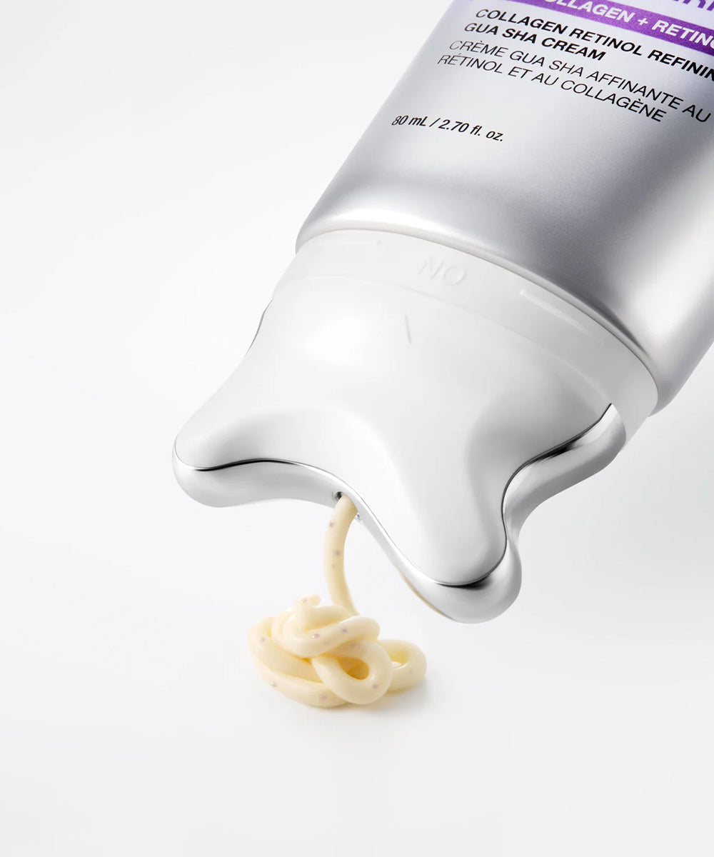 A close-up of a silver cosmetic tube dispensing a pale cream-colored serum or cream onto a clean white surface. The tube has a metallic finish with a white cap and a purple label section at the top.