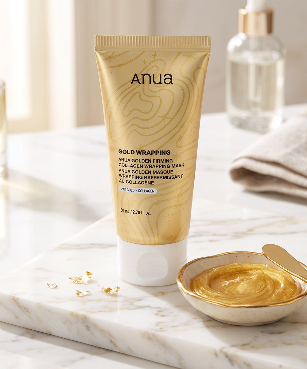 Anua Gold Wrapping Mask tube standing on a white marble tray, with a small bowl of golden cream and gold leaf accents in a bright bathroom-like setting.