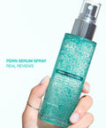 A hand holding the Anua PDRN Serum Spray bottle against a bright white background, featuring the text "PDRN SERUM SPRAY REAL REVIEWS."