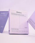 An artistic shot of the white box tilted between two purple mask sachets under soft lavender lighting