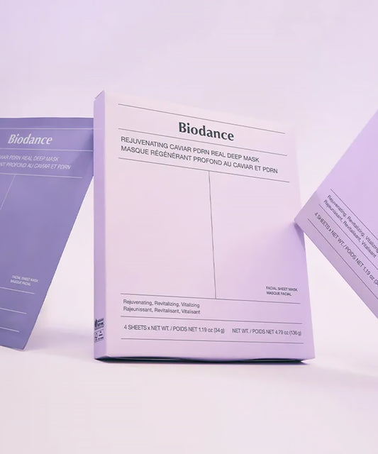 An artistic shot of the white box tilted between two purple mask sachets under soft lavender lighting