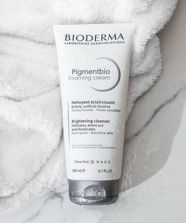 Overhead lifestyle shot of a 200ml tube of Bioderma Pigmentbio Foaming Cream lying on a plush white towel and grey-veined marble surface, featuring water droplets for a refreshing texture