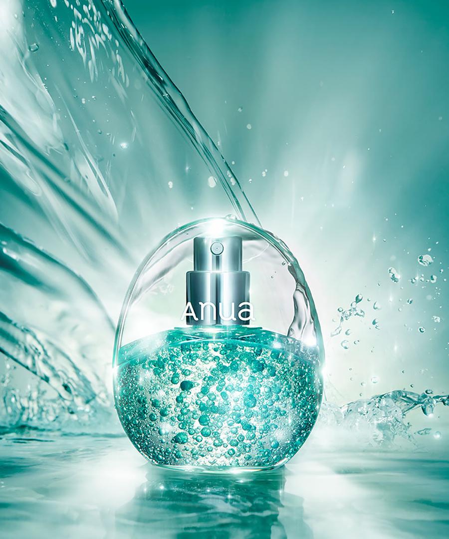 A clear spherical perfume bottle filled with sparkling aqua liquid and silver atomizer, set against a water-splashed teal background. The bottle has the brand name 'anua' printed on the front.