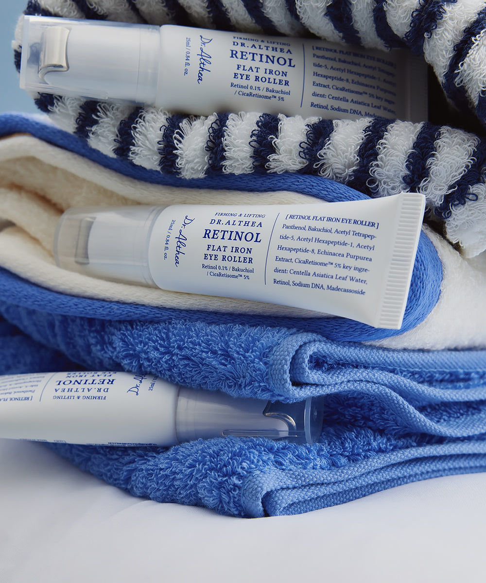 Lifestyle photography of three Dr. Althea Retinol Flat Iron Eye Roller tubes resting on plush blue and white striped bathroom towels, emphasizing a clean and refreshing skincare routine