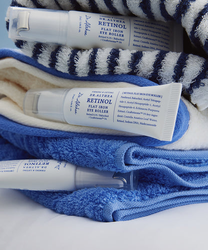 Lifestyle photography of three Dr. Althea Retinol Flat Iron Eye Roller tubes resting on plush blue and white striped bathroom towels, emphasizing a clean and refreshing skincare routine