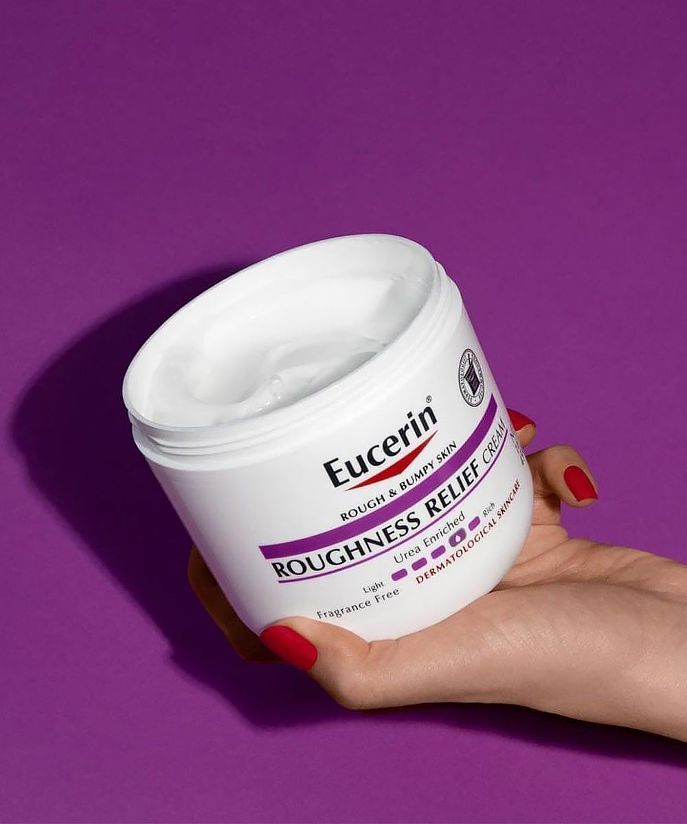 Hand holding a jar of Eucerin Roughness Relief Cream against a purple background