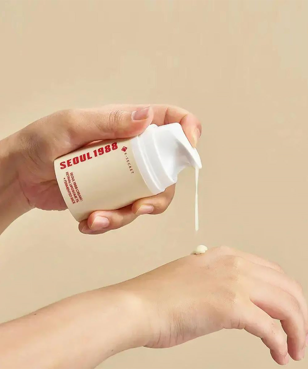 Close-up shot of hands using the K-SECRET Seoul 1988 Cream pump bottle, actively dispensing the beige-colored retinal cream onto the back of another hand against a warm background