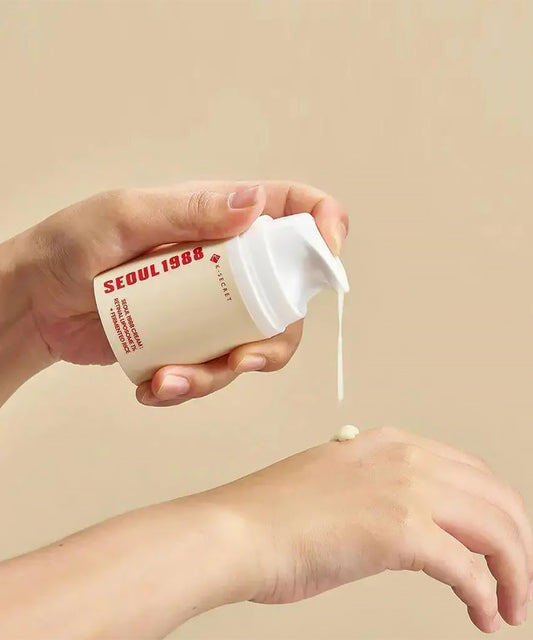 Close-up shot of hands using the K-SECRET Seoul 1988 Cream pump bottle, actively dispensing the beige-colored retinal cream onto the back of another hand against a warm background