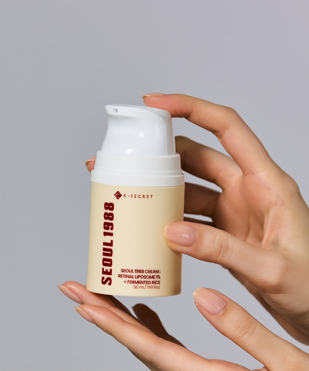 A close-up view of elegant, manicured hands holding the K-SECRET Seoul 1988 Cream pump bottle, with product label details clearly visible against a cool gray background