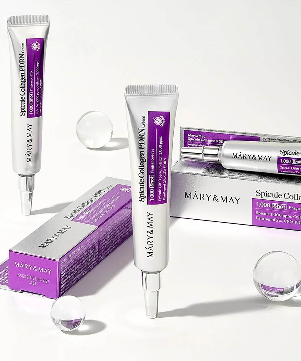 An aesthetic group arrangement of Mary & May Spicule Collagen PDRN Cream tubes and boxes on a white surface with decorative clear spheres and soft lighting