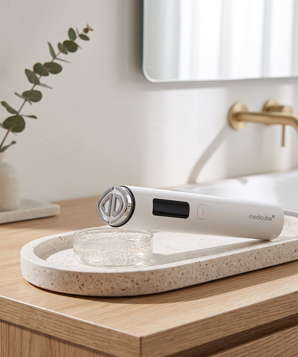 A white handheld skincare device with a digital display and metallic circular head, resting on a speckled beige tray on a light wood vanity. A small glass bowl sits beside it, with a softly blurred bathroom sink, mirror, and brass faucet in the background.
