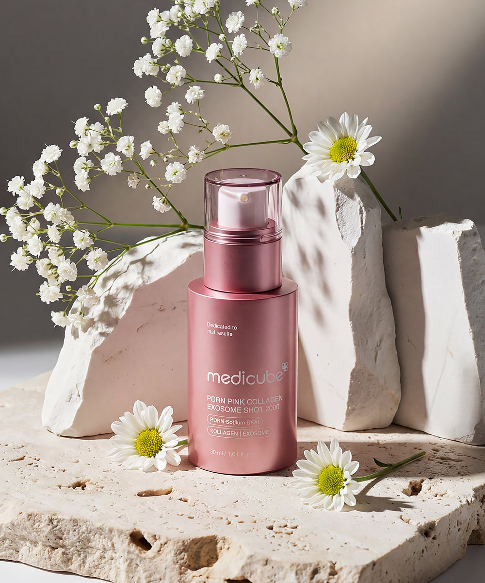 Aesthetic lifestyle shot of the Medicube PDRN Pink Collagen Exosome Shot 2000 placed on a textured stone platform, styled with white daisies and baby's breath flowers