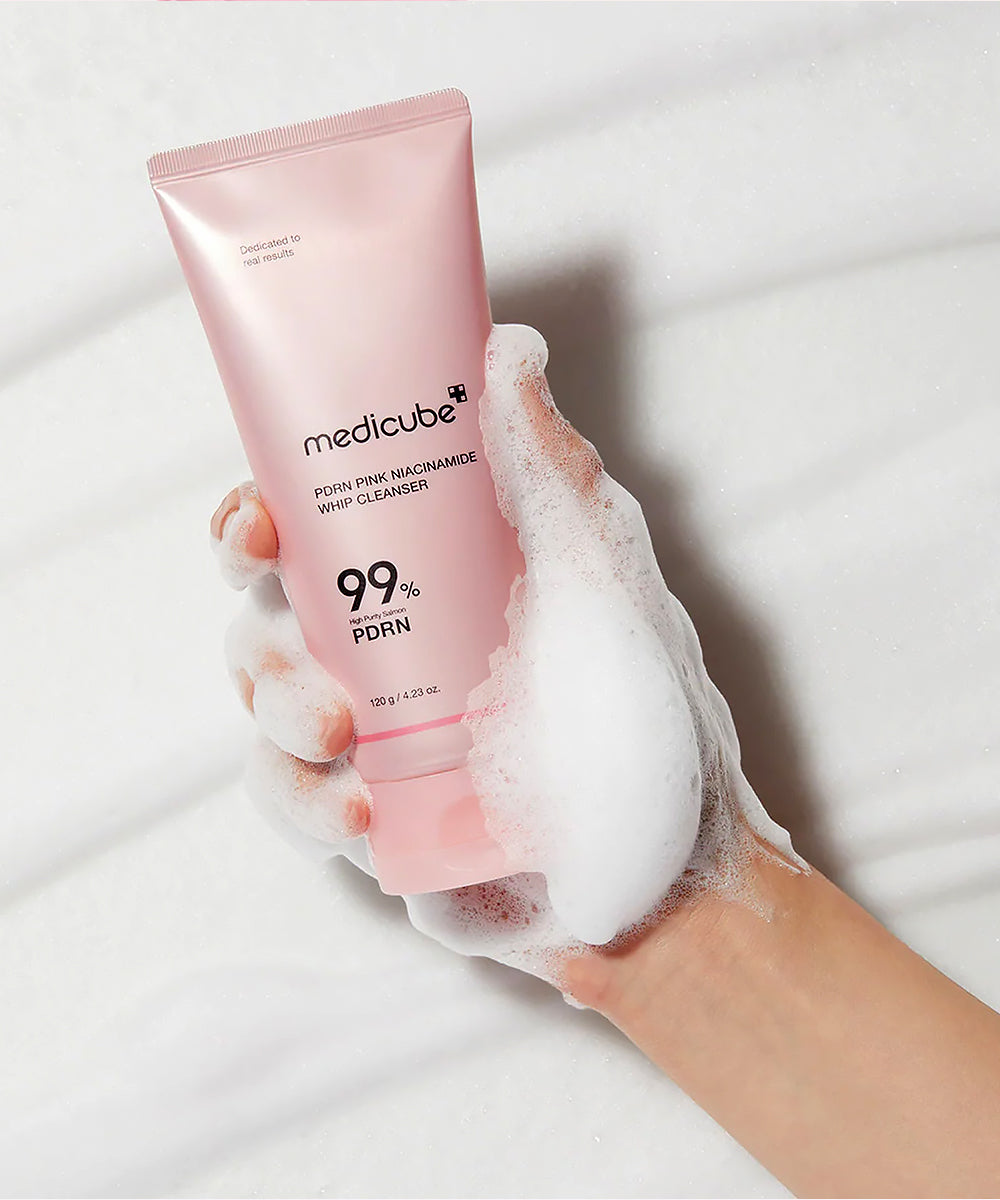 A close-up view of a hand holding the Medicube PDRN Pink Niacinamide Whip Cleanser tube, covered in rich, thick white lather to demonstrate the whipped texture