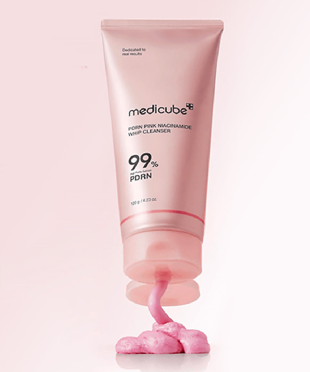 Vertical product shot of the Medicube PDRN Pink Niacinamide Whip Cleanser standing on a pink background, with a dollop of the rich pink cleanser being actively dispensed from the nozzle to showcase its glossy texture