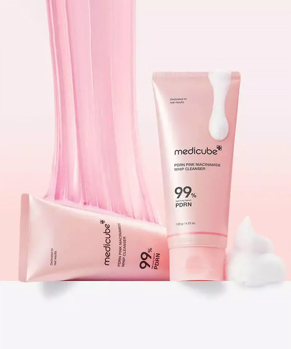 A stylized product composition on a soft pink background, featuring two Medicube PDRN Pink Niacinamide Whip Cleanser tubes—one standing and one lying down—combined with a curtain of pink stretching cleanser and white whipped foam