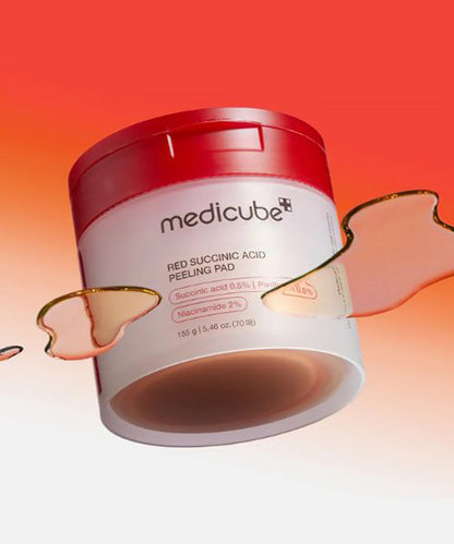 An artistic, angled view of the Medicube Red Succinic Acid Peeling Pad jar, surrounded by floating droplets of amber-colored serum against a vibrant red and orange gradient background