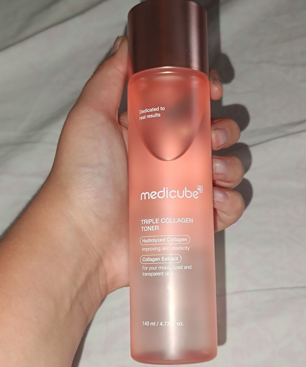 A hand holds the pink Medicube toner bottle, showing its scale and the frosted texture of the packaging against a neutral fabric background