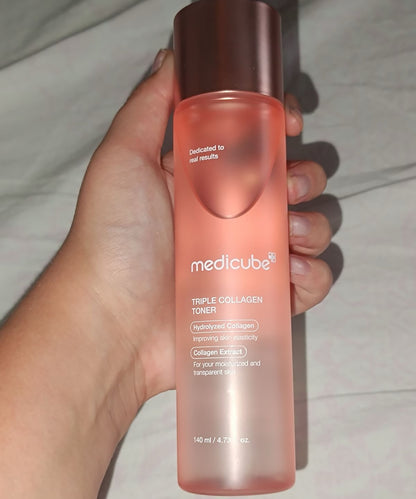 A hand holds the pink Medicube toner bottle, showing its scale and the frosted texture of the packaging against a neutral fabric background
