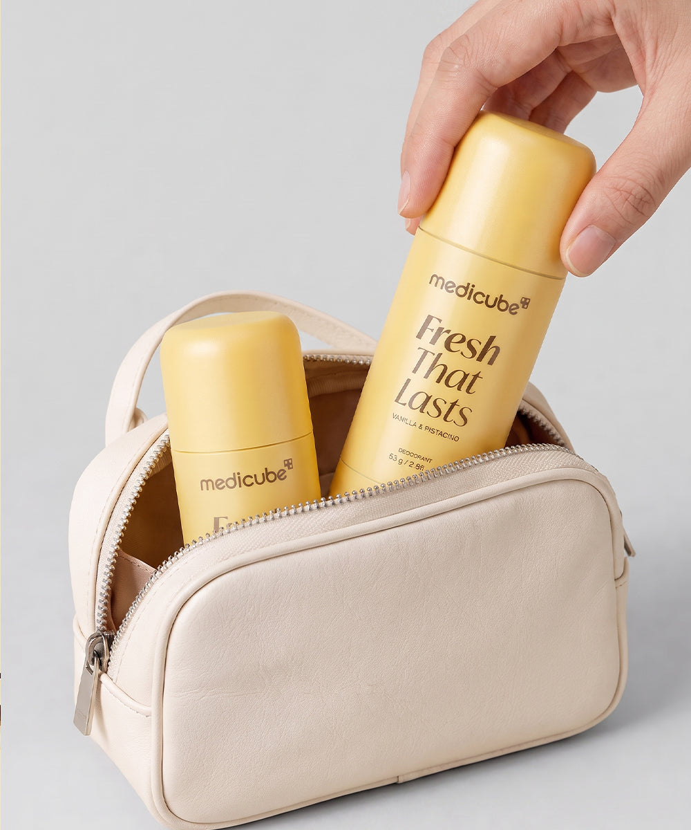 A hand inserts a yellow Medicube Fresh That Lasts travel spray bottle into a beige zippered cosmetic pouch, with a second matching bottle visible inside on a light gray background.