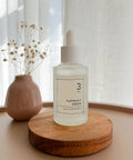 Lifestyle image of the numbuzin No. 3 Skin Softening Serum displayed on a circular wooden coaster next to a small pink vase with dried flowers in a bright, airy setting