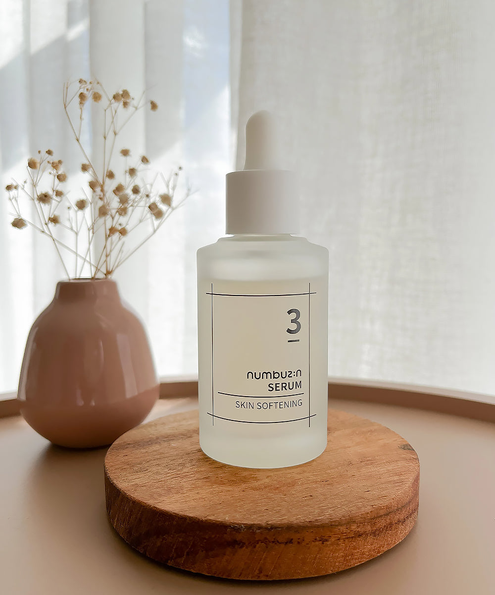 Lifestyle image of the numbuzin No. 3 Skin Softening Serum displayed on a circular wooden coaster next to a small pink vase with dried flowers in a bright, airy setting