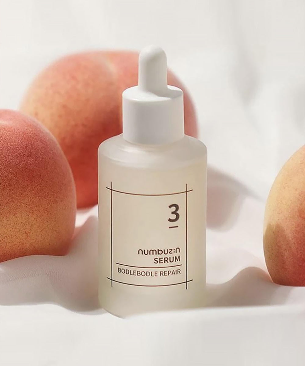 Aesthetic product shot of the numbuzin No. 3 Bodlebode Repair Serum nestled among fresh peaches on a soft, draped white fabric background