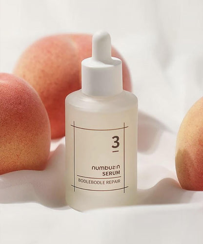 Aesthetic product shot of the numbuzin No. 3 Bodlebode Repair Serum nestled among fresh peaches on a soft, draped white fabric background