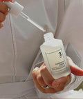 Close-up of the numbuzin No. 3 Bodlebode Repair Serum being held by a person in a white garment, with the glass dropper lifted to show the clear, lightweight texture