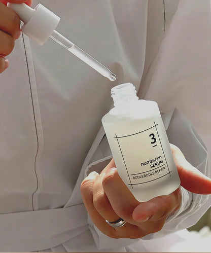 Close-up of the numbuzin No. 3 Bodlebode Repair Serum being held by a person in a white garment, with the glass dropper lifted to show the clear, lightweight texture