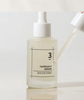 Detailed view of the numbuzin No. 3 Skin Softening Serum with a glass dropper dripping a clear, viscous serum onto a reflective white surface to showcase product consistency