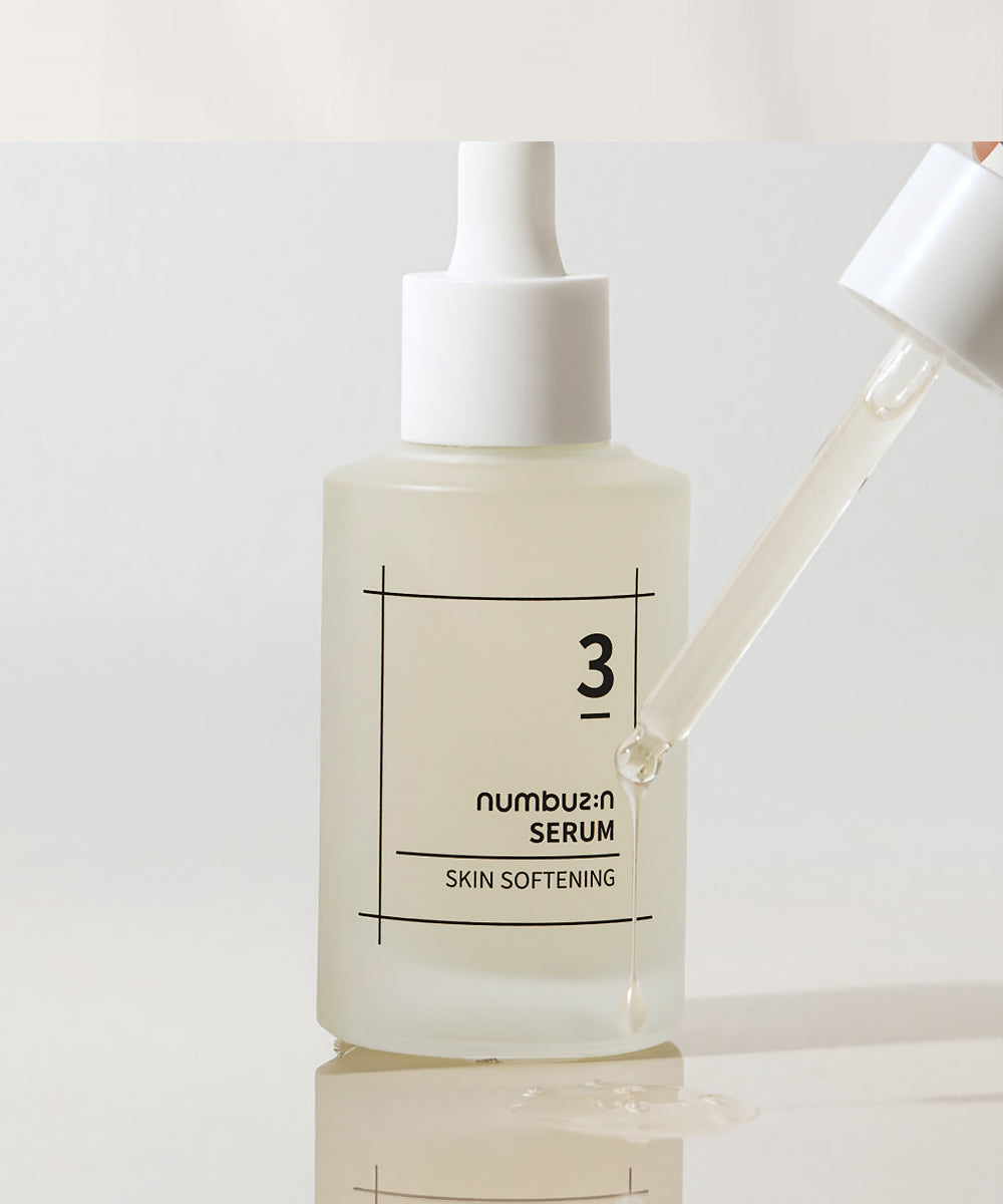 Detailed view of the numbuzin No. 3 Skin Softening Serum with a glass dropper dripping a clear, viscous serum onto a reflective white surface to showcase product consistency