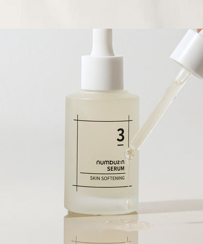 Detailed view of the numbuzin No. 3 Skin Softening Serum with a glass dropper dripping a clear, viscous serum onto a reflective white surface to showcase product consistency