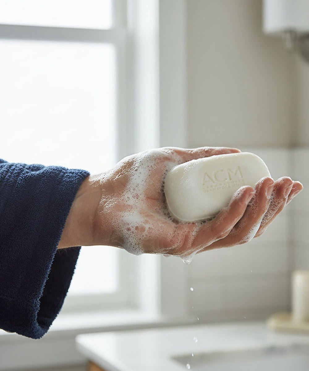 A person’s hand wearing a dark blue terrycloth robe holds the etched ACM soap bar, which is covered in rich white lather, showing the product in use