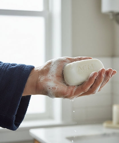A person’s hand wearing a dark blue terrycloth robe holds the etched ACM soap bar, which is covered in rich white lather, showing the product in use