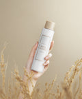 Hand holding a bottle of rice toner with a beige background and dry grass at the bottom.
